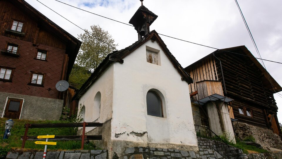 A small white chapel among traditional wooden houses. In the background, trees and a cloudy sky can be seen.