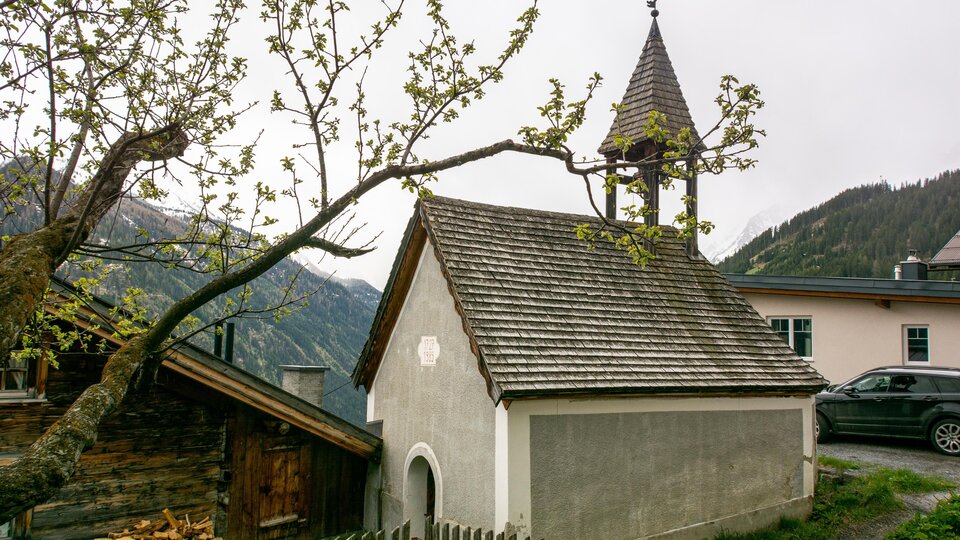 Eine kleine Kapelle mit einem spitzen Dach und einem Kirchturm ist umgeben von Bäumen und Bergen. Im Vordergrund steht ein Holzzaun. | © Bernhard Gruber – Photography