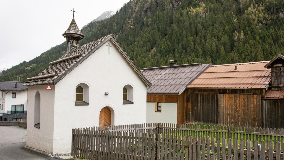 A small, white-painted church with a pointed roof stands in a rural setting. In the background, green mountains and traditional wooden houses can be seen. | © Bernhard Gruber – Photography