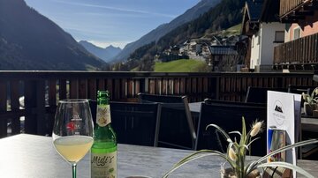 A beautiful table on a terrace with a view of the mountains. On the table, there is a glass with a drink, a bottle, a cup of coffee, and a small plant.