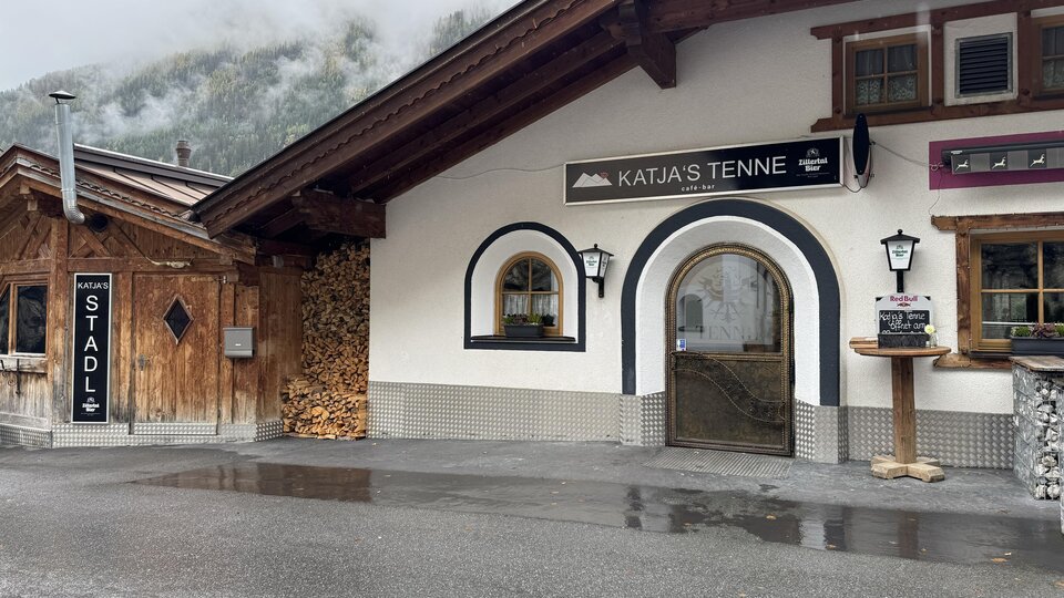 A traditional alpine building with an inviting entrance. Next to the building is a stack of wood and a sign that reads "STADL".