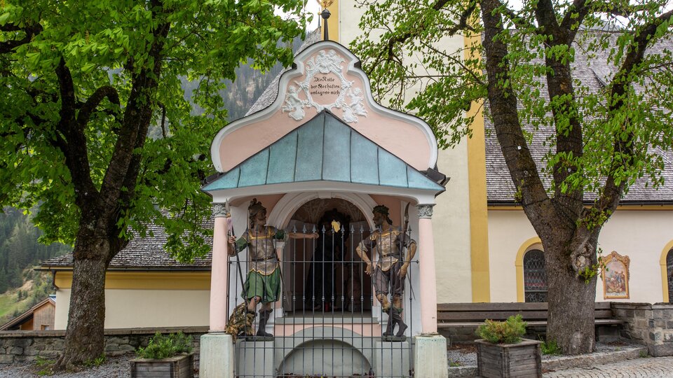 Eine kleine Kapelle mit bunten Figuren und einem grünen Dach. Im Hintergrund stehen große Bäume und ein Gebäude mit gelber Fassade. | © Bernhard Gruber – Photography