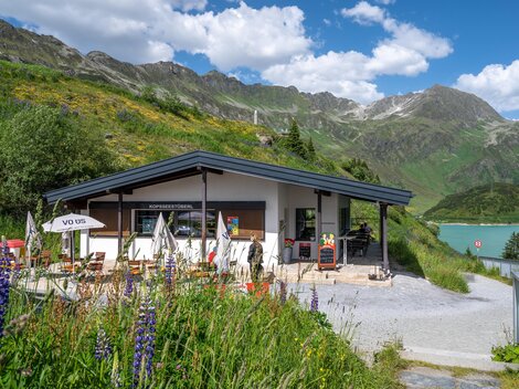 Ein modernes Gebäude am Fuße der Berge, umgeben von grünen Wiesen und bunten Blumen. Im Hintergrund sind majestätische Berge und ein klarer blauer Himmel zu sehen.