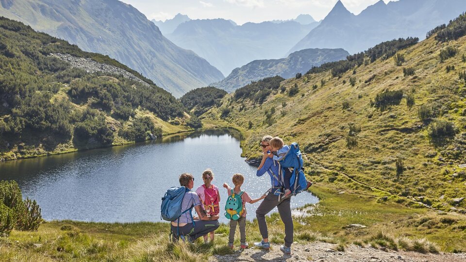 Eine Familie wandert in den Bergen neben einem klaren See. Die Landschaft ist grün und hügelig, umgeben von beeindruckenden Gipfeln.