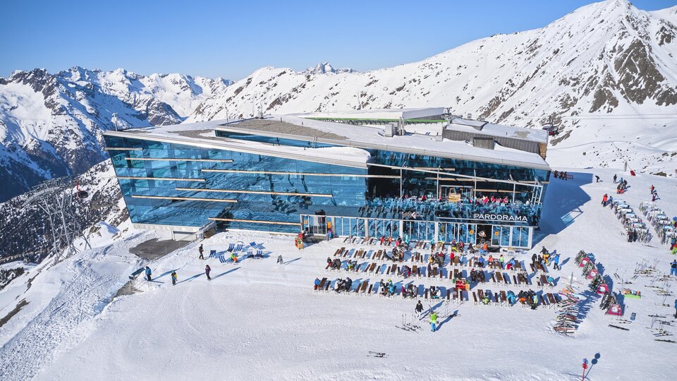 A modern mountain cabin surrounded by snow-covered mountains. Many people are enjoying the sunny day outdoors.