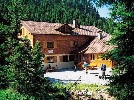 Eine Berghütte umgeben von hohen Tannen. Im Vordergrund sind Wanderer zu sehen, die die Natur genießen.