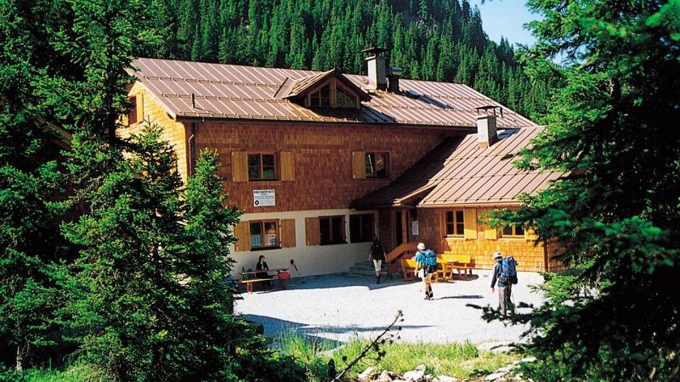 Eine Berghütte umgeben von hohen Tannen. Im Vordergrund sind Wanderer zu sehen, die die Natur genießen.
