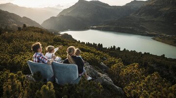 A family is sitting on rocks and looking at a beautiful lake. In the background, mountains and a clear sky can be seen.