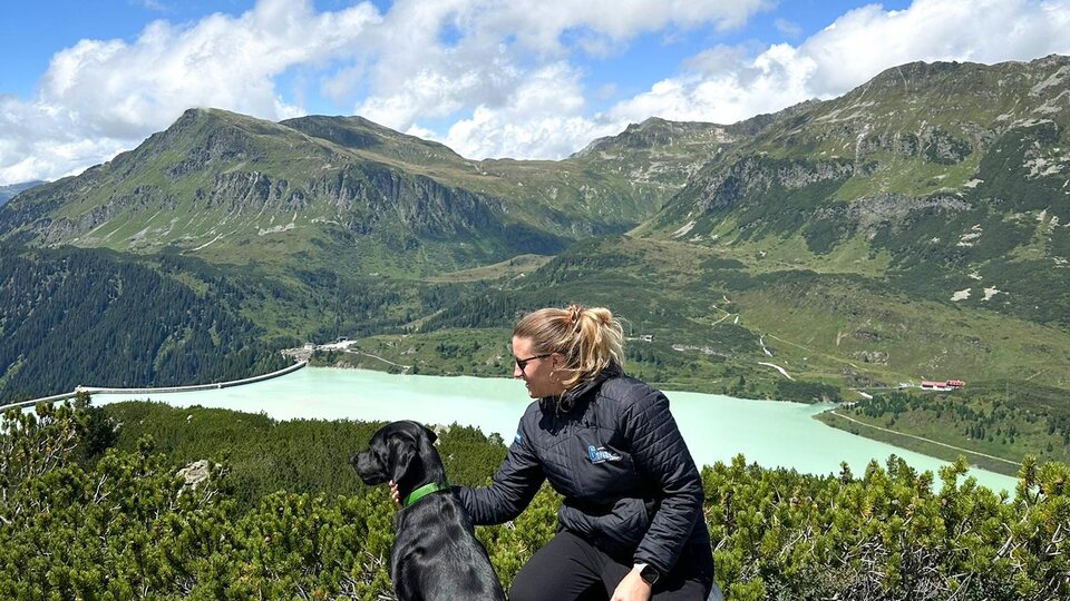 Eine Frau sitzt mit ihrem Hund auf einem Felsen und blickt auf eine grüne Landschaft mit Bergen und einem See. Der Himmel ist blau mit einigen Wolken.