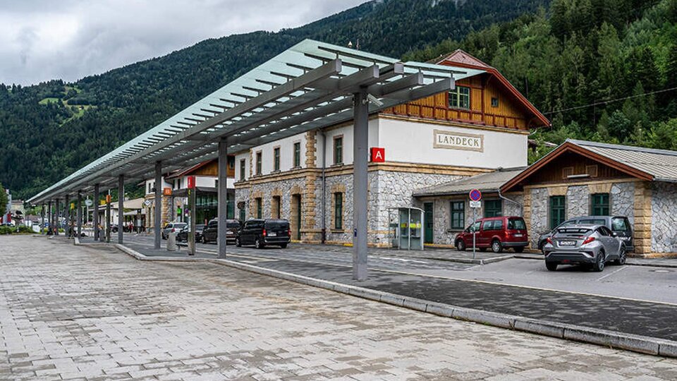 A train station building with traditional architecture and modern roofing. In the background, green mountains and clouds can be seen.