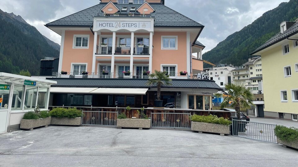 A charming hotel with a bright facade and a balcony on the ground floor. In the background, green mountains and a tranquil atmosphere can be seen.
