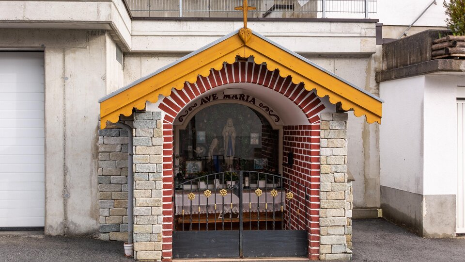 Eine kleine Kapelle mit einem gelben Dach und einem Kreuz. Sie ist aus Stein und Ziegeln gebaut und hat eine geschlossene Gittertür. | © Bernhard Gruber – Photography