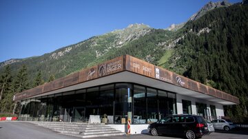 A modern building in the mountains with large windows. The surroundings are surrounded by green forests and mountains.