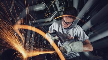 A craftsman is working with a grinding machine and creating sparks. He is wearing safety goggles and work gloves in an industrial environment.