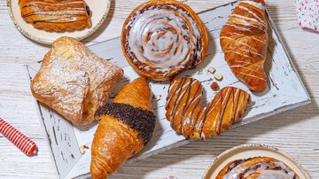 An enticing selection of pastries on a white wooden board. Various items are visible, such as croissants, cinnamon rolls, and filled dough products. | © MPREIS Warenvertriebs GmbH