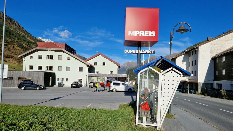 A supermarket named "MPREIS" is visible in a rural setting. In the foreground, there is a bus stop, and in the background, several houses can be seen.