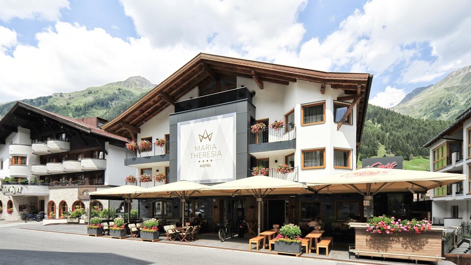 A cozy hotel with an inviting outdoor area and terraces. In the background, green mountains and a blue sky can be seen.