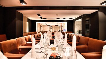 An elegant restaurant space with a long table and cozy armchairs. The tables are festively set with glasses and cutlery.