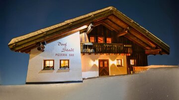 A traditional Alpine house with a wooden facade and an extended roof. It is illuminated and visible against a clear, dark sky.