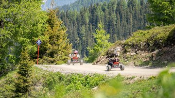 Zwei Personen fahren in roten Karts auf einem schottrigen Weg durch eine grüne Waldlandschaft. Im Hintergrund sind hohe Bäume und Berge zu sehen.
