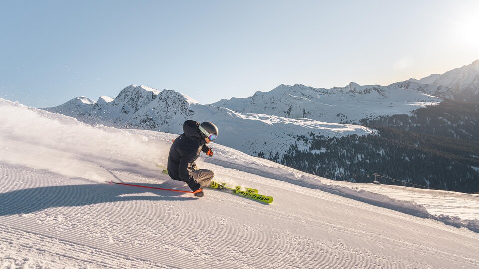 Ein Skifahrer fährt über eine verschneite Piste in den Bergen. Im Hintergrund sind beeindruckende Berglandschaften zu sehen.