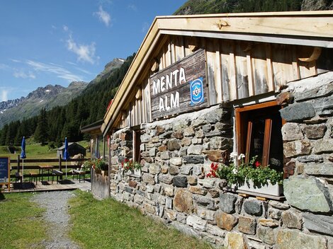 Eine gemütliche Almhütte aus Stein in einer malerischen Berglandschaft. Die Umgebung ist grün, mit Bergen im Hintergrund und blauer Himmel.