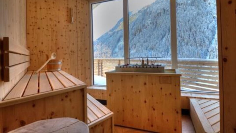 Ein gemütlicher Wellnessbereich aus Holz mit Blick auf schneebedeckte Berge. Das Licht strömt durch große Fenster und schafft eine entspannende Atmosphäre.