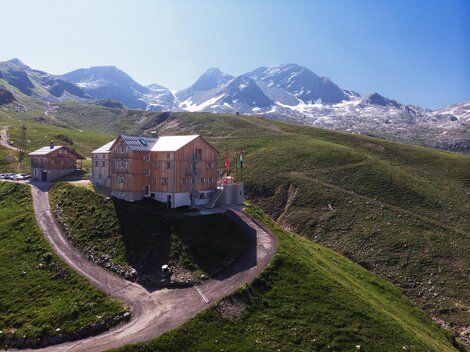 A modern building on a green hill, surrounded by mountains. In the background are snow-capped peaks and a clear blue sky.