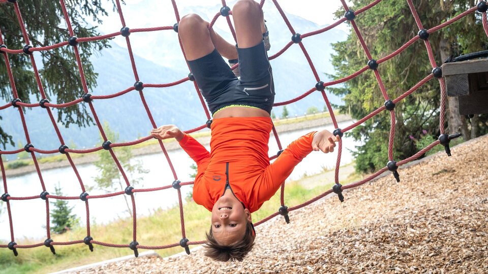 Ein Junge hängt kopfüber in einem roten Kletternetz und blickt fröhlich in die Kamera. Im Hintergrund sind Bäume, ein See und die verschwommene Berglandschaft zu sehen. Das Bild fängt einen spielerischen Moment voller Energie, Mut und Leichtigkeit im alpinen Outdoor-Erlebnis ein.