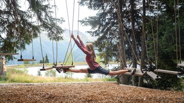 Ein Mädchen balanciert mit ausgestreckten Beinen zwischen zwei schwebenden Holzelementen eines Niederseilgartens. Hinter ihr öffnet sich der Blick auf einen Bergsee und die umliegende alpine Landschaft. Die Szene wirkt sportlich, frei und abenteuerlich und zeigt Naturerlebnis und Geschicklichkeit inmitten der Paznauner Bergwelt.