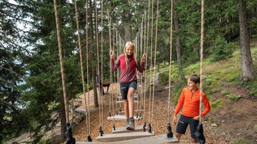 Zwei Kinder erkunden einen Waldseilgarten inmitten dichter Fichten. Ein Mädchen balanciert konzentriert über schwebende Holzbretter, während ein Junge in orangefarbener Sportkleidung daneben steht und lächelnd zu ihr hinüberschaut. Die natürliche Umgebung mit Waldboden, Bäumen und alpiner Kulisse vermittelt Abenteuer, Bewegung und Familienzeit in der Natur.