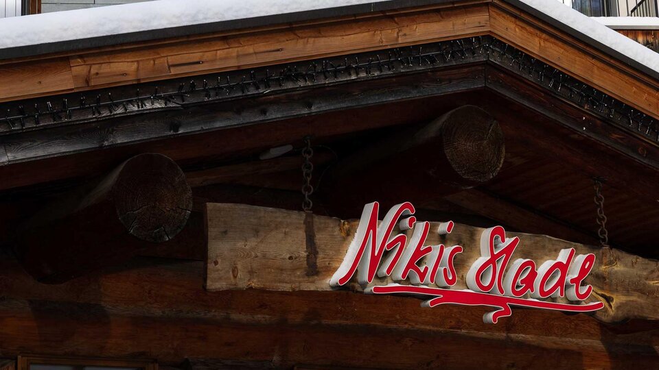 Ein traditionelles Holzgebäude mit einem schneebedeckten Dach. Das Schild mit der Aufschrift "Nikis Stube" ist gut sichtbar.