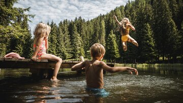 Drei Kinder spielen am Ufer eines Sees. Ein Kind springt fröhlich ins Wasser, während die anderen zuschauen.
