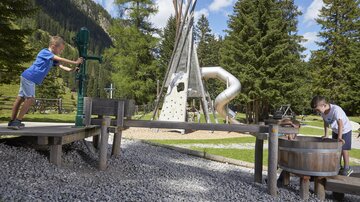 Ein Spielplatz in der Natur mit verschiedenen Spielgeräten. Zwei Kinder spielen aktiv mit einer Wasserpumpe und einem Fass.