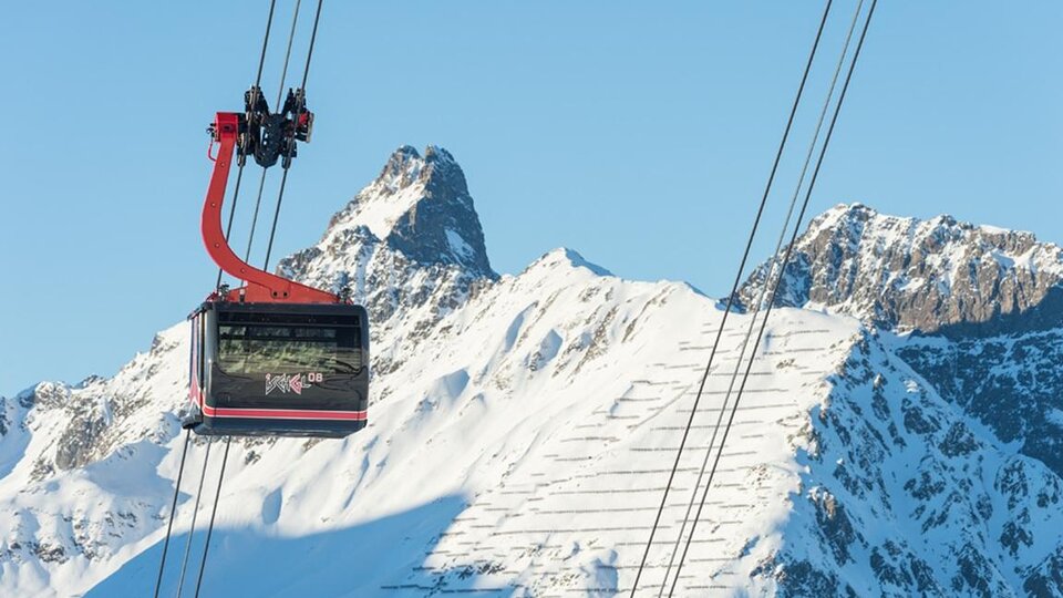 Eine Seilbahn schwebt über schneebedeckte Berge. Der Himmel ist klar und blau.