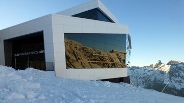 Ein modernes Gebäude in den Bergen, umgeben von Schnee. Die große Fensterfront spiegelt die beeindruckende Berglandschaft wider.