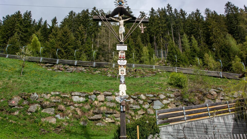 Ein kunstvoller hölzerner Kreuzweg mit verschiedenen Figuren und Symbolen. Im Hintergrund erstreckt sich eine grüne Wiese und bewaldete Hügel. | © Bernhard Gruber – Photography