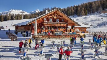 A cozy mountain cabin in the snowy landscape, surrounded by trees. Many people enjoy the winter atmosphere and stand at tables outdoors.