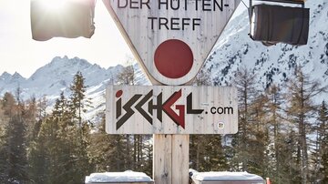 Das Bild zeigt ein Schild für die Paznauner Taja, einen beliebten Treffpunkt in den Bergen. Im Hintergrund sind schneebedeckte Berge und Bäume zu sehen.