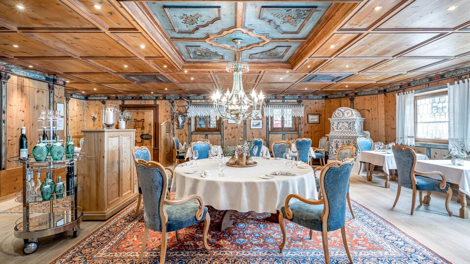Ein eleganter Restaurantbereich mit einem runden Tisch und blauen Stühlen. Das Zimmer ist mit Holzverkleidungen, einem kunstvollen Deckenmuster und einem prächtigen Kronleuchter dekoriert.