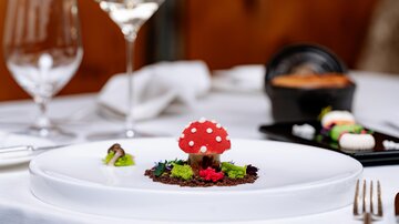 Ein kunstvoll angerichtetes Gericht mit einem roten Pilz aus Schokolade, der auf einem Bett aus buntem Gemüse und Kräutern platziert ist. Im Hintergrund sind Weingläser und weitere Speisen sichtbar.