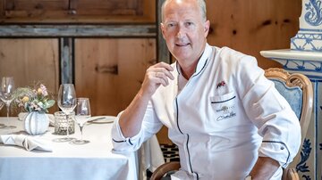 Ein Koch in einer eleganten Restaurantumgebung sitzt an einem Tisch. Der Raum ist gemütlich und stilvoll eingerichtet.