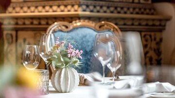 Ein elegantes Tischarrangement mit Gläsern und einer kleinen Blumenvase. Im Hintergrund ist ein kunstvoll verzierten Ofen zu sehen.
