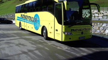 Ein gelber Reisebus mit der Aufschrift „Paznauntaler“ steht auf einem Parkplatz. Im Hintergrund sind grüne Hügel und einige Gebäude sichtbar.