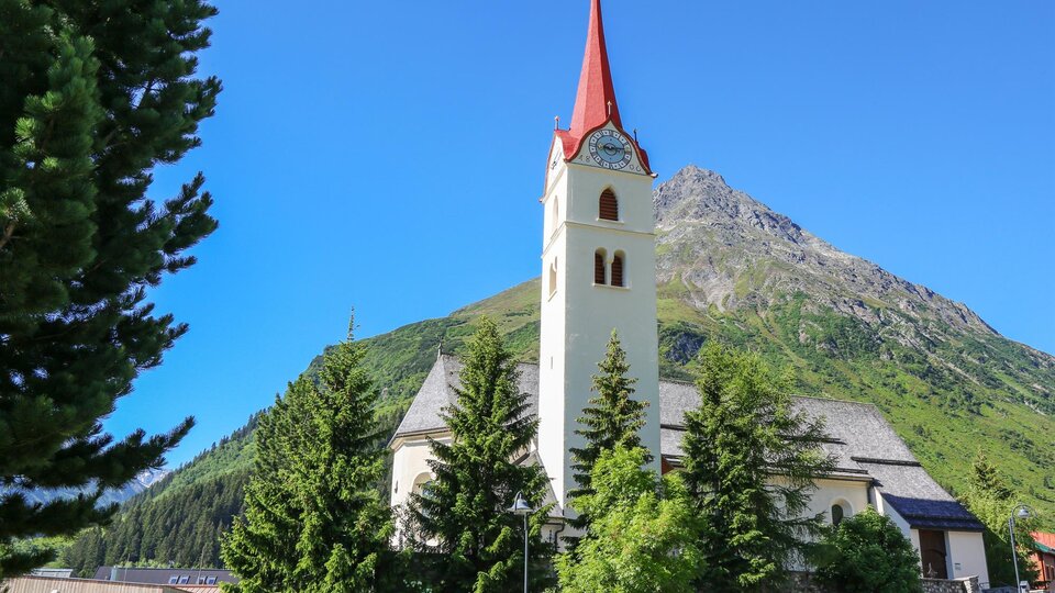 A picturesque church with a red tower, surrounded by green trees and mountains. The sky is clear and blue. | © TVB Paznaun - Ischgl