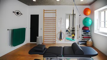 A modern therapy room with a couch and various training equipment. On the wall, there is a climbing wall and exercise balls in different colors.
