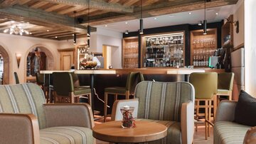 Ein stilvoll eingerichtetes Café mit bequemen Sesseln und einem zentralen Tisch. Im Hintergrund ist eine gut gefüllte Bar sichtbar.