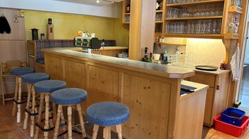Eine gemütliche Bar mit Holzregalen und Gläsern. Es gibt mehrere Hocker mit blauen Sitzkissen und eine einladende Atmosphäre.