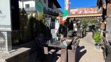 Eine einladende Terrasse eines Restaurants mit Tischen und Stühlen. Im Hintergrund sind grüne Bäume und ein blauer Himmel zu sehen.