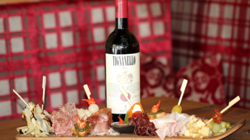 A stylish arrangement of various types of sausage and cheese on a wooden board, surrounded by fresh sides. A bottle of red wine complements this, creating a cozy atmosphere.